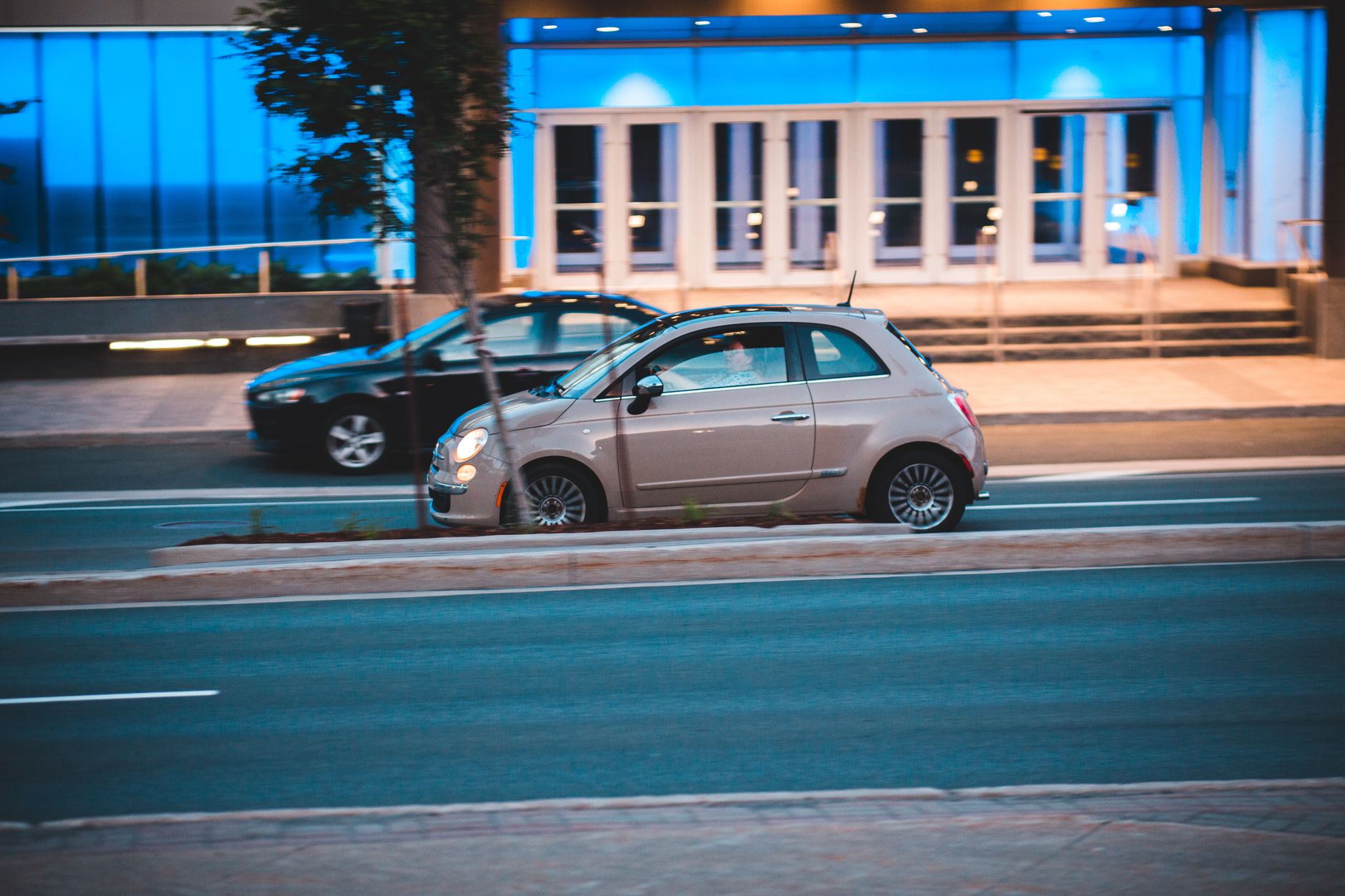 Fiat 500 jadący nocą obok nowoczesnego, niebiesko podświetlonego budynku